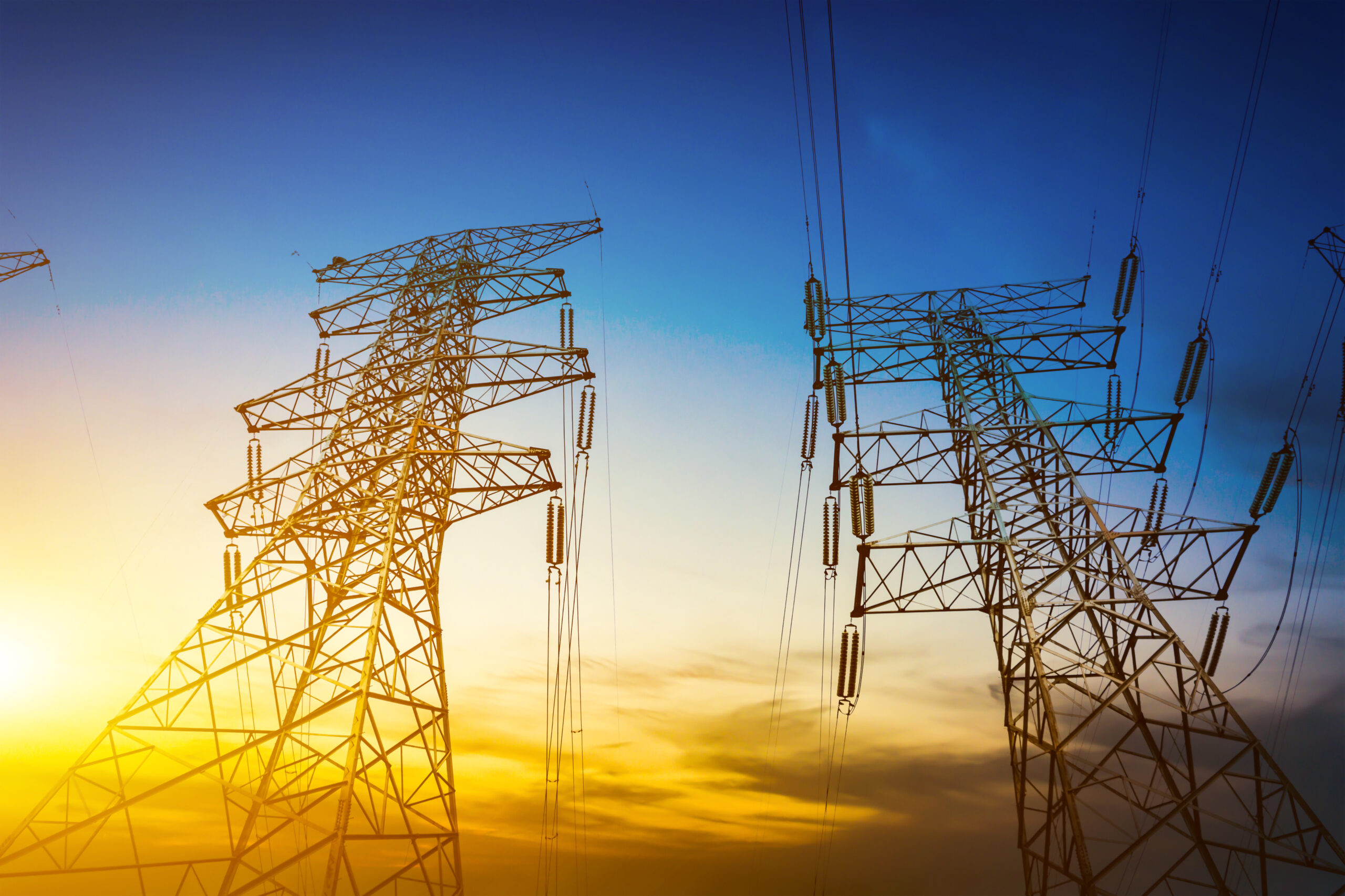 Silhouettes of two large electrical transmission towers against a vibrant sunset sky. The image represents critical power utility infrastructure and evokes the themes of transition, future planning, and financial resilience needed to navigate a decade of disruption.